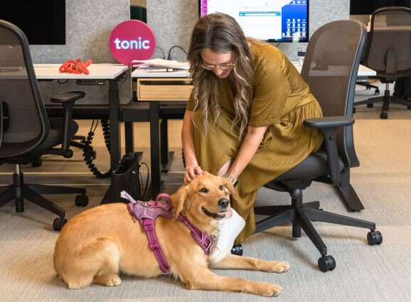 Woman sitting at her desk, reaching down to pet a smiling golden retriever.